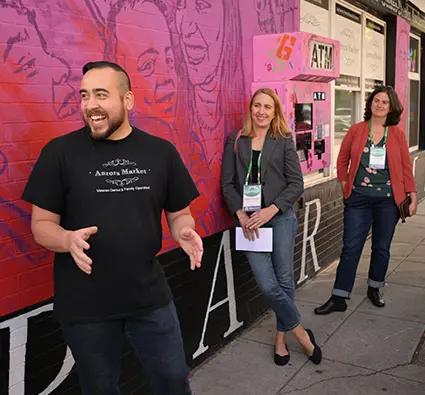 A man in a T-shirt that says "Aurora Market" speaks to a group outside in front of a pink mural of people's faces.