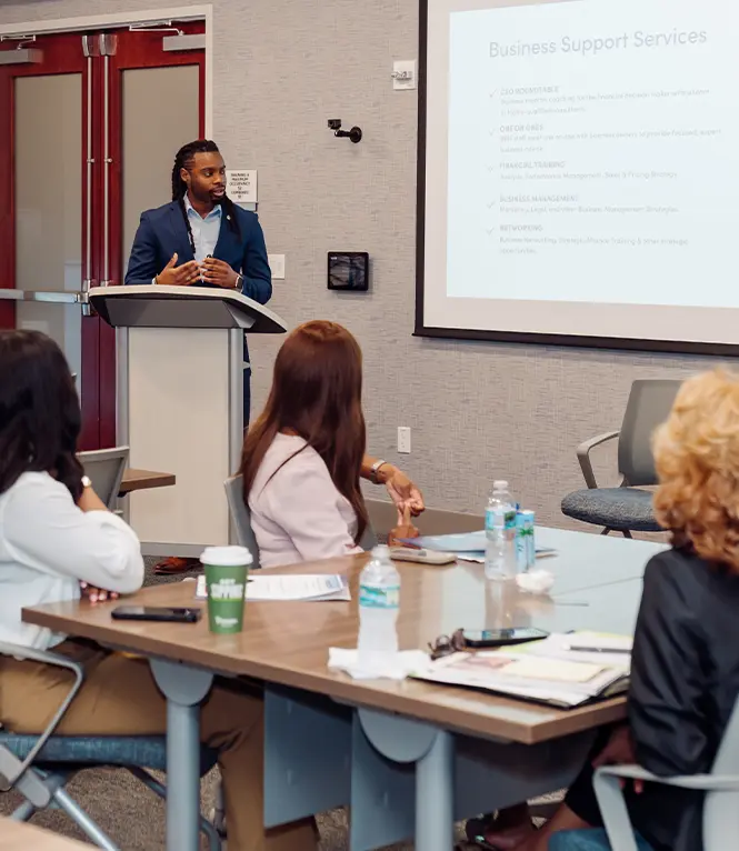 A man stands at a podium at the front of a room and presents slides to a group of people seated at tables.