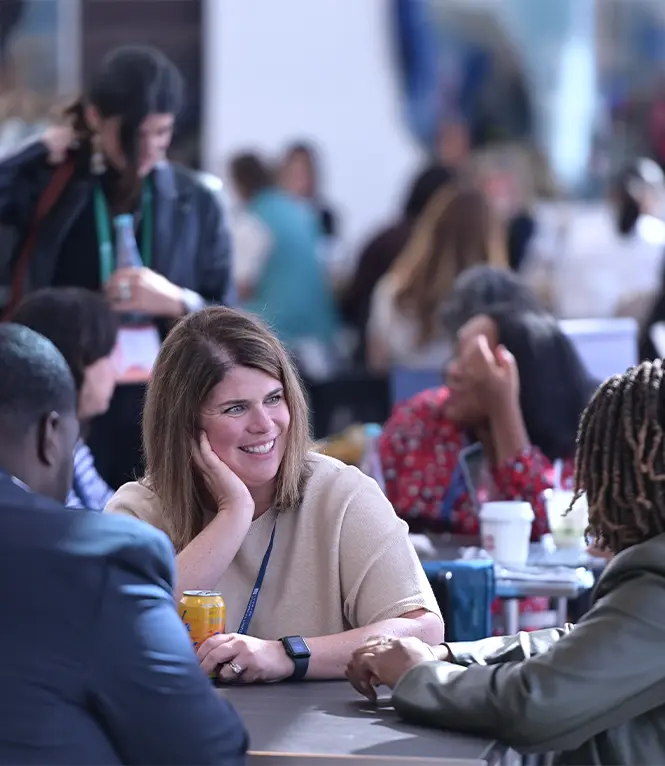 Three people seated at a table in conversation, in an indoor space filled with people. One woman's face is visible and she is smiling.