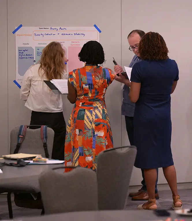 A group of four people in a hotel conference room stand and add notes to a poster taped to the wall.