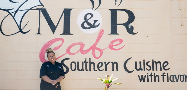 Chef Maria Rumlin standing in front of a wall painted with the M&R Cafe logo