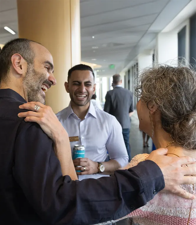 Three people standing in a group, talking and laughing. Two people have their hands on each others' shoulders.