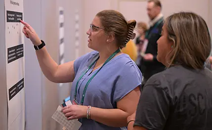 Two women look at a poster on the wall, while one points to the poster.