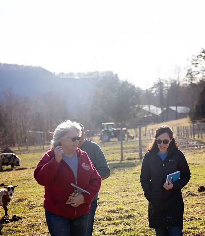 Two women from Mountain Association holding notebooks, walking on a farm with a goat.
