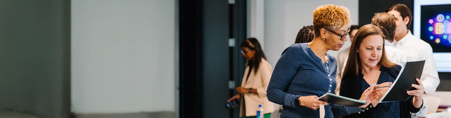 Two women stand in a room of people and look over a notebook together.