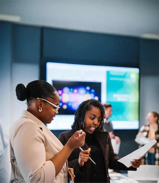 Two women standing indoors, one holding a pen and paper while the other gestures with a pen.
