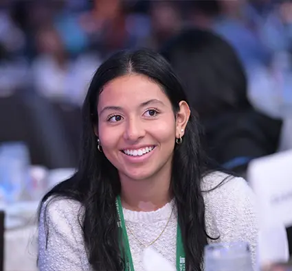 A young woman with a conference badge seated in a room full of people, smiling.